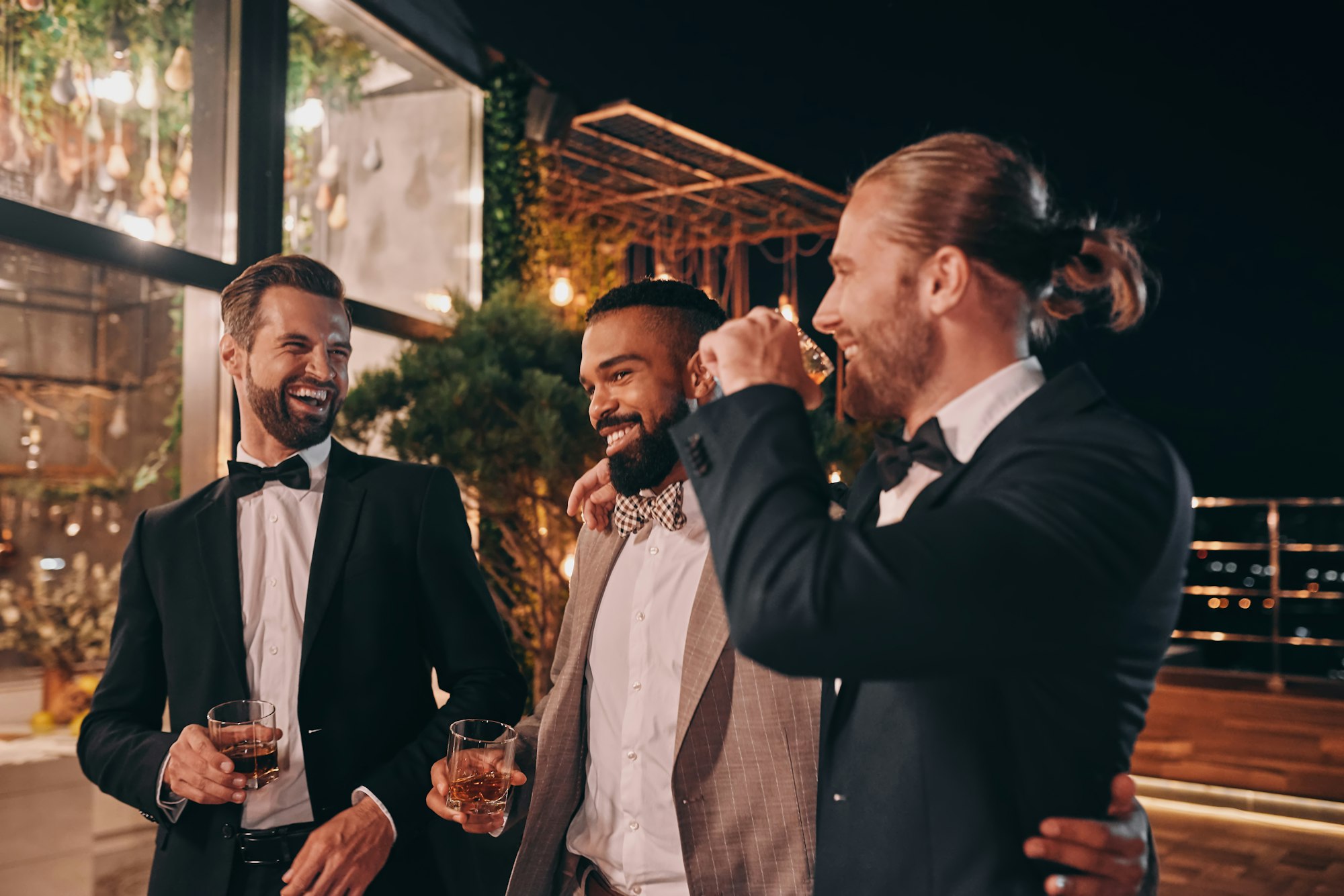 Three happy men in suits holding glasses with whiskey while spending time on party